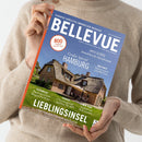 Person in beigefarbenem Strickpullover hält das Immobilienmagazin „BELLEVUE“ mit blauem Titelhintergrund. Auf dem Titelbild ist ein reetgedecktes Backsteinhaus unter blauem Himmel zu sehen. Sichtbare Texte: „EUROPAS GRÖSSTES IMMOBILIEN-MAGAZIN“, „Großes Special HAMBURG“, „LIEBLINGSINSEL – Sylt zwischen Sehnsucht und Realität“, sowie Themen zu Österreich, Spanien, New York und weltweiten Immobilienangeboten.