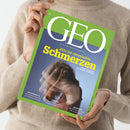 Person in beigefarbenem Strickpullover hält eine Ausgabe des Magazins „GEO“ mit grünem Rand und großem weißem Titelkopf. Das Cover zeigt eine mehrfache, transparente Aufnahme einer Person mit gebeugtem Oberkörper und Händen am Kopf vor blauem Hintergrund. Gelber Titeltext: „Hilfe bei chronischen Schmerzen“, Untertitel: „Neue Therapien für Körper und Seele“, sowie kleinere Texte zu Psychosomatik, Ghana und Indonesien.
