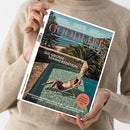 Eine Person in beigefarbenem Pullover hält eine Ausgabe des Magazins „GOODLIFE – Das Magazin für Wohnen & Design, Kultur, Kulinarik, Stil & Reisen“. Auf dem Cover liegt eine Person in gestreiftem Badeanzug auf einer Luftmatratze in einem türkisfarbenen Pool vor mediterraner Landschaft mit Meer und Palmen, darüber die Titelzeile „DIE GROSSE SOMMEREDITION“.