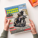 Titelbild des Magazins „Touren Fahrer“ Juli 2026. Eine männlich gelesene Person in Motorradschutzkleidung fährt auf einer weißen Ducati DesertX mit rotem Streifendetail auf kurviger Gebirgsstraße. Schlagzeilen: „Alltagstauglich: Kaufberatung Textiljacken“, „Allzweck in Eleganz“, „Moto Guzzi V7 Sport vs. Triumph Speed Twin 900“, „Tauber-Main, Katalonien, Georgien“. Hände halten das Magazin vor hellem Hintergrund.