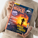 Person mit hellem, langärmligem Strickpullover hält das Magazin „mountain BIKE“ in beiden Händen; Cover mit großem gelbem Titel „Der PREIS ist HEISS“, darunter Text „Das große Duell: Edel-Bike vs. Preiskracher“. Abgebildet ist ein Radfahrender auf einem Berg vor orange-leuchtendem Sonnenuntergang. Weitere Aufschriften: „21 Enduro- & Trail-Reifen im Test“, „Orbea“, „Neu – 16 Seiten Community“, „Shimano XT Di2 im Stresstest“.