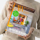 Hellemännliche Person in beigem Strickpullover hält ein Magazin mit orange-weißem Titel „AW Architektur & Wohnen“. Auf dem Cover ist ein modern eingerichtetes Wohnzimmer mit buntem Teppich, gelbem Stuhl, orangem Sessel, Pflanzen und großem Wandbild abgebildet. Sichtbare Texte: „52 Seiten Bad & Wellness“, „Frischer Wind – neue Architektur aus Dänemark“, „Tapeten-Trends – kunstvolle Muster & Texturen“, „Wohnen wie im Urlaub“.