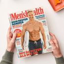 Titelblatt des Magazins „Men’s Health“ mit großem roten Schriftzug oben. Im Mittelpunkt steht ein lächelnder, muskulöser Mann in Badehose, der ein Surfbrett trägt und vor dem Meer steht. Text auf dem Cover: „DEIN BESTER SOMMER“, „67 Tipps für mehr Muskeln und einen flachen Bauch“, „MIT 50 FITTER ALS MIT 35!“, „Einfach genial: Men mit Jamie Oliver“, sowie kleinere Schlagzeilen zu Fitness-, Beziehungs- und Gesundheitsthemen. Hände halten das Heft über hellem Tisch.