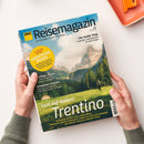 Titelbild des „ADAC Reisemagazin“ mit Berglandschaft, grünem Tal und wandernder Person vor hohen Felsen unter blauem Himmel mit Wolken. Aufschriften: „Lust auf Italien! Trentino“, „City-Guide: Prag“, „Kroatiens Inseln“, „Schweden erleben“ und gelber Kreis mit „14 Seiten Extra Abenteuer-Reisen“. Gehalten von zwei Händen über hellem Untergrund, oranges Objekt oben rechts im Bildrand.
