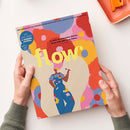 Magazincover „flow“ mit gelbem Schriftzug in der Bildmitte, darunter eine illustrativ dargestellte Person in blauer Latzhose mit bunten Blumenmustern vor abstraktem Hintergrund in Rot‑, Rosa‑, Gelb‑ und Blautönen. Oben der Schriftzug „Das Magazin für persönliche Entwicklung, Kreativität & mehr Ruhe“, rechts Zitat „Je mehr du dein Leben feierst, desto mehr gibt es zu feiern.“ in schwarzer Schrift. Zwei Hände halten das Magazin vor hellem Untergrund.