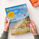 Titelblatt des „ADAC Reisemagazin“ mit blauem Himmel, Strand und Dünenlandschaft an der Ostseeküste, auf einem Hausdach weht die dänische Flagge, im Vordergrund gehen eine Person mit weißem Kleid und eine Person mit hellem Hemd am Strand entlang; Haupttitel „Sommerglück in Dänemark“, weitere Texte: „Urlaub ohne Hitze“, „Auf nach Paris!“, „Ernten neu erleben“, „Traumreise: Kanada“. Zwei Hände halten die Zeitschrift vor hellem Hintergrund.
