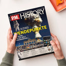 Titelbild des Magazins „P.M. HISTORY“ mit dunkelblauem Hintergrund, großem gelben Schriftzug „WENDEPUNKTE“ und Unterzeile „Von Hiroshima bis zum iPhone – 75 Jahre, die die Welt veränderten“. Sichtbare Motive: Atompilz, Astronaut, Brandenburger Tor mit Menschenmenge, iPhone mit App-Symbolen. Unten Textkästen mit Überschriften „SAMT & SEIDE“, „HALLO?!“ und „WM-FIEBER“. Zwei Hände halten das Heft über einem weißen Tisch neben einer orangefarbenen Schale.