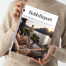Person in beigem Strickpullover hält eine Ausgabe des Magazins „Robb Report“ mit dem Titel „BODY & MIND ISSUE“. Auf dem Cover sitzen und stehen zwei hell gekleidete Personen auf einer Terrasse mit Blick auf einen See und Berge bei Sonnenuntergang. Sichtbare Aufschrift: „Luxus der Regeneration – Die besten Kliniken, Routinen und Investitionen für ein langes, gesundes Leben und was die Wissenschaft wirklich empfiehlt.“