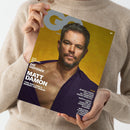 Titelblatt der Zeitschrift „GQ Germany“, gehalten von einer Person in beigem Strickpullover. Auf gelbem Hintergrund ist ein männlicher Schauspieler mit kurzem grauem Haar und leicht geöffnetem dunkelviolettem Hemd abgebildet. Textaufschriften: „Das Making-of Die Odyssee mit Matt Damon, Tom Holland & Robert Pattinson“ sowie weitere Themen zu Jeremy Pope, Pride-Special, Watch Edit, Uhren, WM 2026 und Spielerstars.