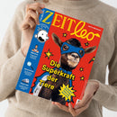 Eine Person in beigefarbenem Pullover hält das Magazin „ZEIT Leo“ mit rotem Titelhintergrund. Auf dem Cover eine Ziege mit blauer Superheldenmaske und Umhang, umgeben von gelben Sternen und dem Schriftzug „Die Superkraft der Tiere“. Am blauen Seitenstreifen links sind ein gezeichneter Kinderkopf, Fotos von Personen sowie Texte zu Themen wie „WM-Stars“, „Spurensuche“ und „Tempo 300“ sichtbar.
