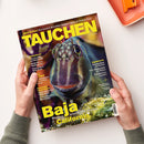 Titelbild der Zeitschrift „Tauchen“, Ausgabe Juli 2026, gehalten von zwei Händen vor hellem Hintergrund. Im Zentrum ein close-up eines bunten Meeresfisches mit markanten Augen und filigranen Hautfortsätzen. Gelbe Schrift mit Artikeltiteln wie „Baja – Mexikos artenreicher Westen“, „Equipment“, „Inselcheck!“ und „Exklusive Leserreisen“. Farben dominieren in Blau-, Grün- und Gelbtönen mit glänzender Oberfläche des Covers.
