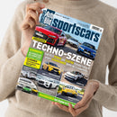 Eine Person ohne sichtbares Gesicht hält ein schräg ausgerichtetes, glänzendes Magazin mit dem Titel „Auto Bild sportscars“ vor einem beigen Strickpullover. Das Cover zeigt mehrere rote, blaue, gelbe, weiße und schwarze Sportwagen sowie die Texte „VERGLEICHSTEST“, „TECHNOLOGIE...“, „Audi RS 5 HYBRID und Elektro Hyundai Ioniq 6 N gegen Verbrenner BMW M3 Competition“ und „GEWINNSPIEL“.