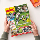 Titelblatt der Zeitschrift „selbst – Das Do-it-yourself-Magazin“, gehalten von zwei Händen über hellem Tisch. Auf dem Cover: ein Mann mit mehreren Mährobotern auf einer Wiese, Schlagzeilen u. a. „Smarte Mäher ohne Draht“, „Wasser im Garten nutzen“, „Kleine Solaranlage – so sparen Sie Strom“, dazu Fotos von Gartenarbeiten, Werkzeugen und Heimwerkerprojekten, farbige Textfelder in Gelb, Blau, Rot und Weiß.
