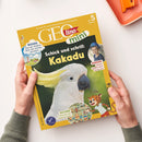 Kinderzeitschrift „GEOlino mini“ mit gelbem Titelrand, großem Foto eines weißen Kakadus mit gelbem Federkamm auf grünem Hintergrund, Schriftzug „Schick und schrill: Kakadu“. Oben rechts Hinweis „ab 5 Jahren“, unten runde Bildausschnitte mit einem lachenden Kind, der Aufschrift „KINDER Kylian gräbt eine Burg aus“ sowie „SUCHEN Erkunde die PUPPENSTUBE“. Zwei Hände halten das Heft auf hellem Tisch neben orangefarbener Schale.