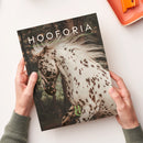 Zwei Hände in grünen Strickärmeln halten ein Magazin mit dem Titel „HOOFORIA“. Das Cover zeigt ein weißes Pferd mit dunklen Flecken in Bewegung, im Hintergrund ein unscharfer Wald. Unterhalb des Titels steht der Untertext „NATURE · ELEGANCE · ATHENE · EQUESTRIAN“, unten mittig ein grünes Logo mit den Buchstaben „H“ und „C“. Rechts oben liegt eine orange Schale auf einer weißen Tischfläche.