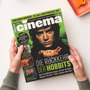 Magazin mit dem Titel „cinema“ in weiß-grüner Schrift auf grün-schwarzem Hintergrund, gehalten von zwei Händen über hellem Tisch; auf dem Cover eine männliche Person mit lockigen Haaren, die einen goldenen Ring auf den geöffneten Handflächen hält; Textaufschriften u. a. „DIE RÜCKKEHR DER HOBBITS“, „IM CHECK!“, „EXKLUSIV-REPORT: 30 Jahre INDEPENDENCE DAY“ sowie mehrere Filmabbildungen und Untertitel zu Streaming- und Kino-Themen.