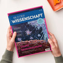 Zeitschrift „BILDER DER WISSENSCHAFT“ mit pinkem Rand und blauem Titelbereich, schräg von zwei Händen einer nicht erkennbaren Person gehalten; das Cover zeigt leuchtende Quallen und die Überschrift „Unser Immunsystem“. Sichtbare Texte: „Im Fokus: Monde mit Tiefenmeeren: Verbergen sich dort exotische Lebensformen?“, „Bestäuber: Populäre Honigbienen, unterschätzte Wespen“ und „Smart Home“.