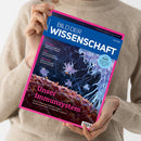 Person mit beigefarbenem Strickpullover hält eine schräg fotografierte Zeitschrift mit pinkem Rand und blauem Titelbereich. Lesbare Texte: „BILD DER WISSENSCHAFT“, „Unser Immunsystem“, „Bestäuber“, „Limes“ und „Smart Home“; das Titelbild zeigt mikroskopisch wirkende helle Zellstrukturen auf dunklem Hintergrund. Das Geschlecht der Person ist nicht erkennbar.