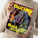 Magazin „TAUCHEN“ mit gelbem Schriftzug auf dem Titelblatt, gehalten von einer hell gekleideten Person vor neutralem Hintergrund. Das Cover zeigt ein Unterwasserfoto eines Fisches in Nahaufnahme mit grünlich-violetter Färbung. Sichtbare Überschriften: „Baja – Mexikos artenreicher Westen, California“, Themen über Inselcheck, Inland-Abenteuer und Tauchequipment in gelber und weißer Schrift auf dunklem Hintergrund.