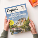 Magazin „Capital“, Ausgabe 05 Mai 2025, mit blau-weißem Titelblatt und der Aufschrift „Der große Immobilien-Kompass 2025“. Darunter Bild einer weißen Stadtvilla mit rotem Ziegeldach und Boot auf einem Fluss, umgeben von Bäumen. Gelber Kreis mit Text „Trump-Crash – Aktien, Anleihen, Gold: So schützen Sie Ihr Depot“. Gehalten von zwei Händen über hellem Untergrund mit orangefarbener Ablageschale oben rechts.