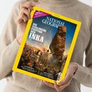 Person in beiger Strickjacke hält eine Ausgabe des Magazins „National Geographic“ mit gelbem Rand und der Titelseite „Die verlorene Festung der Inka“. Das Titelbild zeigt eine historische Szene mit mehreren Figuren in traditioneller Kleidung vor einem hohen Felsen unter blauem Himmel. Sichtbare Textelemente: „Juli 2026“, „Rettendes Reptil“, „Amerika feiert“ und „World’s Biggest Analog Project“.