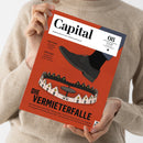 Eine Person ohne sichtbares Gesicht hält ein rot-orangefarbenes Magazin mit weißem Titelbereich und schwarzer Typografie. Das Cover trägt „Capital“ und „WIRTSCHAFT IST GESELLSCHAFT“ sowie die Texte „CHINA-GEFAHR“, „AKTIVE FONDS“ und „DIE VERMIETERFALLE“; abgebildet sind ein schwarzer Schuh und ein Ring aus Häusern.