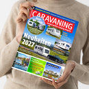 Person in beigefarbenem Strickpullover hält mit beiden Händen ein schräg fotografiertes Magazin. Das farbige Cover trägt die Aufschrift „CARAVANING – Europas großes Camping-Magazin“ sowie „Neuheiten 2027“ und zeigt mehrere Wohnwagen, ein Zugfahrzeug, Landschaftsaufnahmen und weitere Texte wie „Schutzbriefe für Camper“, „Adria Alpina“ und „Tabbert Vivaldi“.
