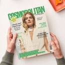 Schräg von oben fotografierte „COSMOPOLITAN“-Zeitschrift auf weißer Fläche, von zwei Händen gehalten; oben rechts liegt ein orangefarbenes Notizbuch. Das Cover zeigt eine blonde Frau im weißen Oberteil sowie die Texte „THE MONEY ISSUE“, „FUN. FEARLESS. FEMALE.“, „Meghan Fahy“, „Charis-ma? Charis-me!“, „FULL FACE LOOKS“ und „Girls just wanna have funds!“.