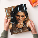 Titelbild der Zeitschrift „ELLE“ vom Juli 2026, gehalten von zwei Händen über hellem Untergrund. Auf dem Cover eine weibliche Person mit dunklem Haar, bronze-braunem Oberteil und sandfarbenem Hintergrund. Textelemente: „MAGIC GLOW – Mini-Touch-Up, Mega Effekt“, „GREECE, MY LOVE! – Das Lebensgefühl, der Look, die Locations“, „HOLLYWOOD GLAM“, Namen: „Zendaya“, „Anne Hathaway“, „Charlize Theron“, „Lupita Nyong’o“.