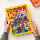 Magazin „National Geographic Kids“ mit gelbem Rand und rotem Titelhintergrund, mittig ein großes Foto einer Eule mit weit geöffnetem Schnabel, flankiert von kleineren Tierbildern und Texten wie „EULEN: Mit großen Augen durch die Nacht!“, „ORCAS“, „PHARAONEN“, „DINOSAURIER“ sowie Hinweis auf „2 starke Poster“; gehalten von zwei Händen über weißem Tisch mit orangefarbenem Tablett.