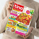 Person in beigefarbenem Strickpullover hält das Magazin „tina Koch & Back-Ideen“ mit rotem Titelkopf. Auf dem Cover sind mehrere Speisen zu sehen, darunter ein Teller mit Hähnchen in Tomatensoße und Basilikum, Heidelbeer-Schnitten, ein Omelett-Wrap und Desserts. Sichtbare Schrift: „Appetit auf Italien“, „Fix aufgetischt!“, „Süße Erfrischung! Heidelbeer­schnitten“ und „Extra – Ihr Heft zum Raushnehmen“.
