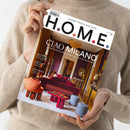 Eine hell gekleidete Person hält das Magazin „H.O.M.E.“ mit der Titelseite „CIAO MILANO – Das Beste vom Salone del Mobile und der Milan Design Week“ in beiden Händen; die Ausgabe zeigt ein modern eingerichtetes Wohnambiente mit rotem Sofa, runden Hockern, glänzendem Tisch und goldfarbenen Wänden, dazu kleinere Titeltexte wie „Neue Möbel“, „EuroCucina“ und „Yacht-Special“ auf weißem Magazindeckblatt.
