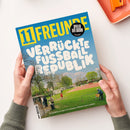 Titelbild der Fußballzeitschrift „11 FREUNDE“ mit gelbem Logo auf blauem Hintergrund und weißer Überschrift „VERRÜCKTE FUSSBALLREPUBLIK“. Abgebildet ist ein Sportplatz mit grünem Rasen, roter Laufbahn, mehreren Personen beim Fußballspielen und Zuschauenden neben dem Spielfeld; unten der Text „Eine Reise zu den kuriosesten Plätzen des Landes“. Zwei Hände halten das Magazin über einer weißen Tischfläche.