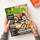 Titelbild der Zeitschrift „bike“, Ausgabe 08/2026, mit grünem Logo oben. Eine maskuline Person mit weißem Helm, rotem Flanellhemd und beiger Hose fährt ein schwarzes Mountainbike im Wald, leicht angehoben auf dem Vorderrad. Aufschriften: „26 Zoll lebt“, „Exklusive Handmade-Bikes“, „Eurobike“, „High-Flow-Ventile“ und gelbes Feld mit „BIKE-PACKING: Traumtour Tessin“. Hände halten das Magazin über hellem Hintergrund mit orangefarbener Ablage.