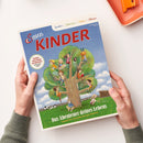 Titelblatt der Kinderzeitschrift „Servus Kinder“ mit großer Illustration eines Baumes, in dem mehrere Kinder unterschiedlichen Alters spielen, lesen und klettern; grüne Wiese mit Kuh und Zaun im Hintergrund unter blauem Himmel. Aufschrift oben: „Spielen | Staunen | Rätseln | Wissen“, unten: „Das Abenteuer deines Lebens – Tausend Ideen für große und kleine Entdecker“.
