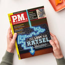 Person mit grüner Strickjacke hält eine Ausgabe des Magazins „P.M. Das Wissensmagazin“ mit gelbem Rand und rotem Titelbalken; Haupttitel: „Die Lust am Rätsel“ über einem schwarzen Labyrinth mit leuchtend blauer Pfeilgrafik, dazu Texte über „Digitale Forensik“, „Lunar Cruiser“ und „Goldrausch“, oben rechts Abbildung zweier Vögel und Schriftfeld „5000 Euro Belohnung für den Geistervogel“.