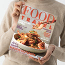 Person in beigefarbenem Strickpullover hält das Magazin „Food and Travel – Deutsch“ mit großem roten Titeltext. Auf dem Cover befindet sich eine Nahaufnahme eines Tellers mit gebratenen Garnelen, garniert mit Kräutern und knusprigen Gewürzen, darüber ein Garnelenstück in Essstäbchen. Sichtbare Schlagzeilen: „Spektakuläre Spas“, „Asiatische Küche“, „Rezepte von der Donau“, „Berlin“, „Vietnam“, „München“ und „Afrikas Küsten Foodie-Hotspots“.