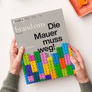 Magazin „brand eins“ mit grauem Titelblatt, schwarzer Überschrift „Die Mauer muss weg!“ und Unterzeile „Modular denken“, oben links Logo „brand eins“ mit Schriftzug „Das geht!“. Unterer Bildbereich zeigt eine Mauer aus bunten, aufgeschichteten Kunststoffbausteinen in Blau, Grün, Gelb, Orange, Rosa und Lila; zwei Hände in grünen Ärmeln halten das Heft über hellem Hintergrund.
