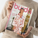 Person in beigefarbenem Strickpullover hält das Magazin „burda style“ mit rosa Schriftzug und heller Titelseite in den Händen; darauf eine weibliche Person in weißem Kleid mit roten floralen Mustern und Texten wie „LUST AUF SONNE!“, „SUNKISSED“, „EXTRA FÜR IHN“ sowie „Brigitte Bardot – Iconic Pattern“, vor verschwommenem Hintergrund mit Kakteen.