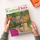 Titelblatt der Zeitschrift „GartenFlora“ vom August 2026, gehalten von zwei Händen über hellem Untergrund. Abgebildet ist ein sommerlicher Garten mit Topfpflanzen in verschiedenen Farben vor einer Steinmauer und zwei Holzstühlen. Textelemente: „Unsere Top 10 der pflegeleichten Kübelpflanzen“, „Lieblingsplätze im Garten und auf der Terrasse“, „Aromatisch – sonnen­gereifte Pfirsiche“, sowie Themenhinweise in einem pinken Kreis unten links.