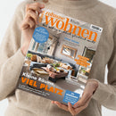 Helle Zeitschrift mit dem Titel „zuhause wohnen – Wie Wohnen wirklich glücklich macht“, gehalten von einer Person in beigem Strickpullover. Auf dem Cover ist ein modernes Wohnzimmer mit grauem Sofa, Holztisch, beigen Kissen und grünen Pflanzen zu sehen. Textaussagen u. a.: „Kleine Räume, VIEL PLATZ“, „Lust auf Süden“, „Kühle Träume“, „Zaubern mit Tapeten“ und „Zu gewinnen“.