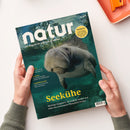 Magazin „natur“ mit Titelbild eines unter Wasser schwimmenden Manatis in grünlich-blauer Umgebung; gelbe Überschrift „Seekühe“ und Untertitel „Mächtige Säugetiere, die Seegras weiden und dabei das Unterwasser-Ökosystem formen“, weiße und gelbe Textbestandteile zu Themen wie „Himalaya“, „Amphibienarche“ und „Arten“. Gehalten von zwei Händen über hellem Untergrund mit orangener Schale im Hintergrund.