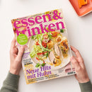 Kochzeitschrift „essen & trinken“ mit dem Titel „essen & trinken“, schräg von oben auf weißer Fläche fotografiert und von zwei Händen mit grauen Ärmeln gehalten. Das Cover zeigt Salat, Gemüse, Geflügel und Garnelen sowie die Texte „Aus Liebe zum Kochen“, „Saucen-Schule 3. Teil: So geht Jus“, „Neue Hits mit Huhn“ und „Italien für Genießer“.