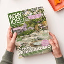 Titelblatt der Zeitschrift „Wohnidee“ mit grün-weißem Schriftzug, gehalten von zwei Händen über hellem Hintergrund. Das Cover zeigt einen dekorierten Gartentisch mit grünem Geschirr, Glasvasen und Blumen unter hängenden Lampen. Sichtbare Texte: „Grüne Wohlfühldeko“, „Hitzeschutz – So wird der Garten fit für neue Wetterlagen“, „Kleine Fläche, viel Sommer!“ und „Design ganz nachhaltig“.