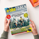Titelblatt des Magazins „e-bike TOUREN MAGAZIN“ in Blau, Gelb und Weiß mit Aufschrift „Die schönste Zeit des Jahres! 20 Traumtouren für Genießer“. Abgebildet sind eine weibliche und eine männliche Person mit Fahrradhelm auf E-Bikes vor Nadelbäumen. Weitere Texte: „45 Ideen für den nächsten Ausflug“, „Märchenhafte Mittelgebirge“, „Marktcheck E-Bike-Motoren“, „Extra neue Helfer für E-Biker“. Hände halten das Magazin über heller Tischfläche.