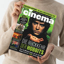 Person in beigem Strickpullover hält ein Magazin mit grün-schwarzem Titelblatt „Cinema“ in beiden Händen; Cover zeigt eine männliche Filmfigur mit lockigem Haar und ernstem Blick, die in ihren Händen einen goldenen Ring hält. Textausschnitte auf dem Heft: „DIE RÜCKKEHR DER HOBBITS“, „Alles zum Mittelerde-Sequel“, „IM CHECK!“, „EXKLUSIV-REPORT: 30 Jahre INDEPENDENCE DAY“, sowie weitere Filmtitel und Bildvorschauen am linken Rand.