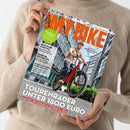 Person in beigem Pullover hält ein Magazin mit dem Titel „MY BIKE“. Auf dem Cover fährt eine männliche Person mit Helm und beiger Jacke ein rotes Fahrrad durch eine städtische Umgebung mit Glasfassaden. Sichtbare Textelemente: „TOURENRÄDER UNTER 1800 EURO“, „Diamant. Decathlon & Stevens im Test“, „NEWCOMER AVENTON“, „FAHRRAD-RUCKSÄCKE“, „PANORAMA-TOUR AUF DER SCHWEIZ“ sowie „BLINKER AM FAHRRAD-CHECK & RECHTSLAGE“.