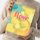 Weibliche Person im beigefarbenen Strickpullover hält ein schräg ausgerichtetes Magazin mit türkis-gelbem Cover, gelben Kreisformen, weißen Linien und kleinem Figurenmotiv. Sichtbare Texte: „flow“, „Das Magazin für persönliche Entwicklung, Kreativität & mehr Raum“ und „Do what you love and be happy“.