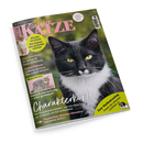 Schräg fotografiertes Titelblatt des Katzenmagazins „Geliebte KATZE“, August 2026, auf schwarzem Hintergrund. Ein großformatiges schwarz-weißes Katzenporträt mit gelben Augen dominiert das Cover; links stehen ein helles Kätzchen und ein kleines grau getigertes Katzenfoto. Sichtbare Texte: „Europas beliebtestes Katzenmagazin“, „Aktuelle Forschung“, „Experten-Tipps“, „Pinkelt-Pannen“, „Charakterkopf“ und „Zum Weltkatzentag“.