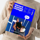Weibliche Person im cremefarbenen Strickpullover hält ein schräg fotografiertes, blaues „Harvard Business manager“-Magazin. Das Cover zeigt zwei Männer, einer mit weißem Headset, sowie die Texte „Wie Game Boy und Crocs zu Ikonen wurden“, „Warum Chatbots umgarnt“, „Zahlen sich immersive Erlebnisse aus?“, „Wann es Zeit ist, Prioritäten neu zu setzen“, „Nö.“, „Jetzt gib schon auf…“ und „Verhandeln am Limit“.